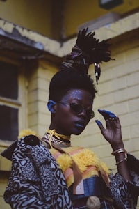 a woman with dreadlocks and a yellow jacket standing in front of a brick wall